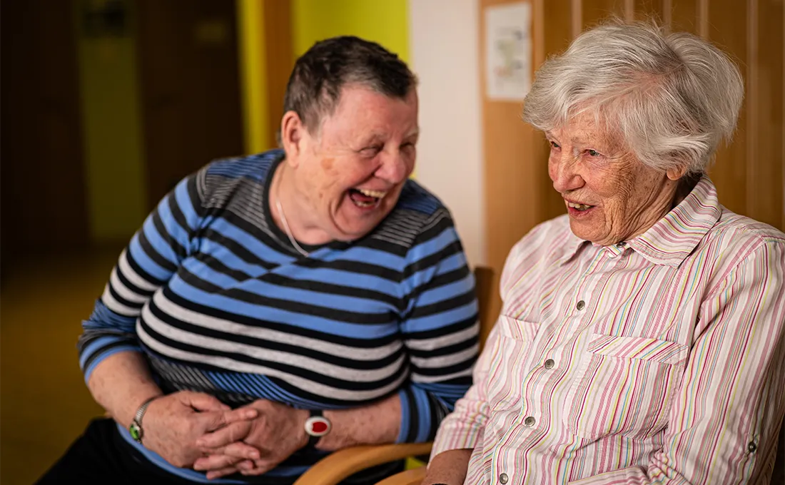 Zwei Seniorinnen sitzen lachend nebeneinander im Gespräch